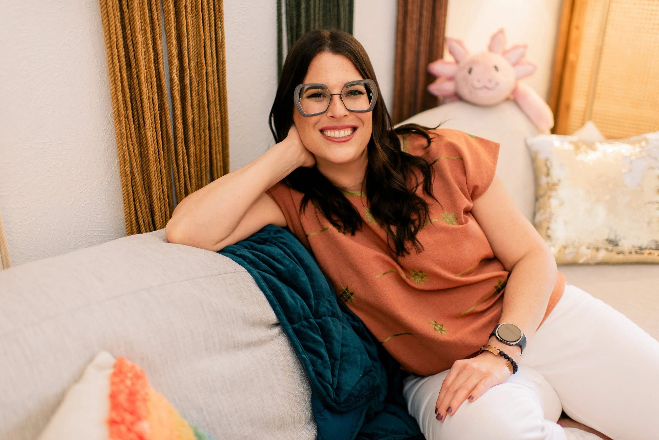 Meet your therapist, Joann Chevaillier, LPC-Associate. Joann has long dark hair and glasses and she's smiling warmly, sitting on a beige couch with colorful pillows and an axolotl stuffed animal.