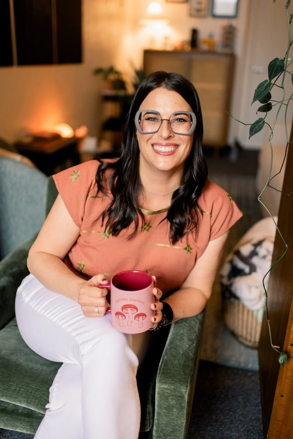 A woman with black hair, glasses, and a peach-colored shirt with star patterns, smiling and sitting on a green armchair, holding a pink mug with mushroom illustrations and the text 'Mushroom Hunter' in a cozy, warmly lit room.
