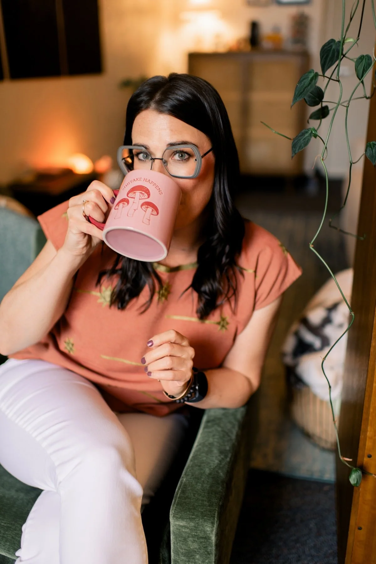 Woman with black hair and glasses sitting on a green couch, drinking from a pink mug with mushroom illustrations, indoors with warm lighting, plants, and wooden furniture in the background.