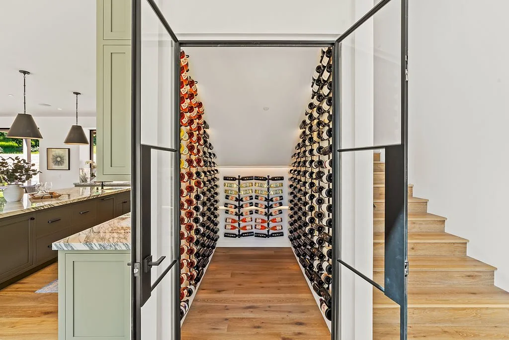 A walk-in wine cellar with wine bottles stored on black metal racks along the walls, with a small rack at the back for additional bottles, and an open glass door leading to a modern kitchen.