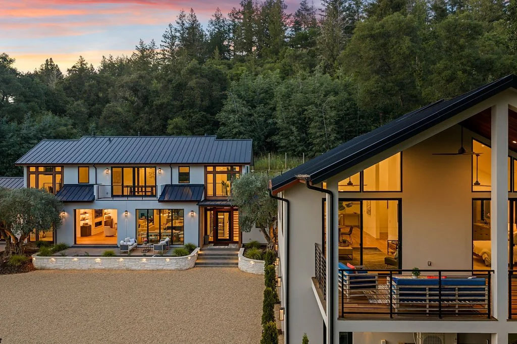 Two modern houses with large glass windows and porches, illuminated from inside, situated on a gravel clearing in front of a forest during sunset.