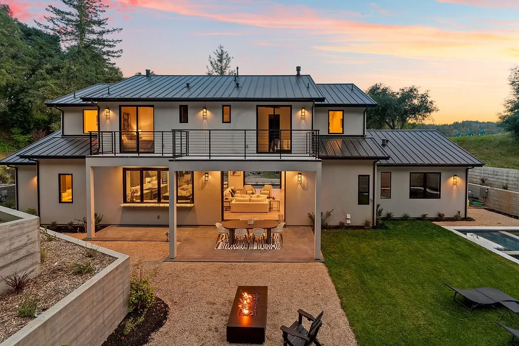 Modern two-story house with a metal roof, illuminated windows and outdoor lights, overlooking a landscaped yard with a fire pit and a small swimming pool at sunset.