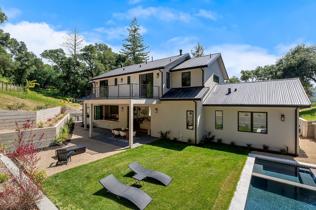 Modern two-story house with a metal roof, balcony, and a backyard with lounge chairs and a swimming pool, surrounded by trees and a partly cloudy sky.