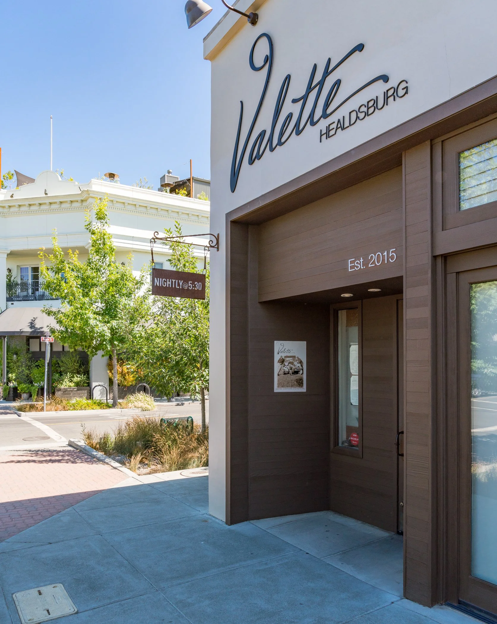 Exterior view of Valette restaurant in Healdsburg, established in 2015, with clear view of its sign on a white wall, and a small window with a poster inside. There is a tree in front and adjacent buildings in the background under a clear blue sky.