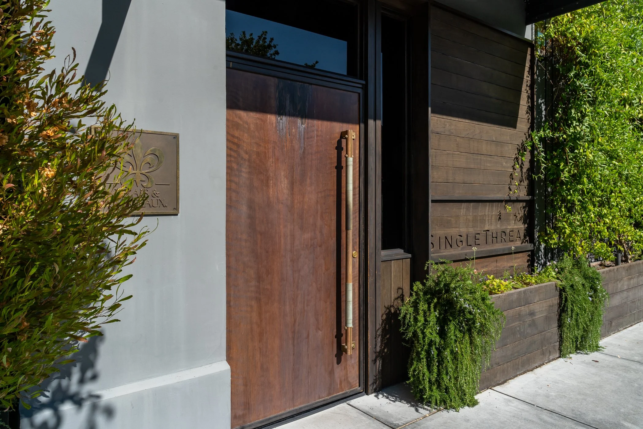 Modern building entrance with a wooden door, metal handle, and a sign on the wall. Green bushes along the building and sidewalk in bright sunlight.