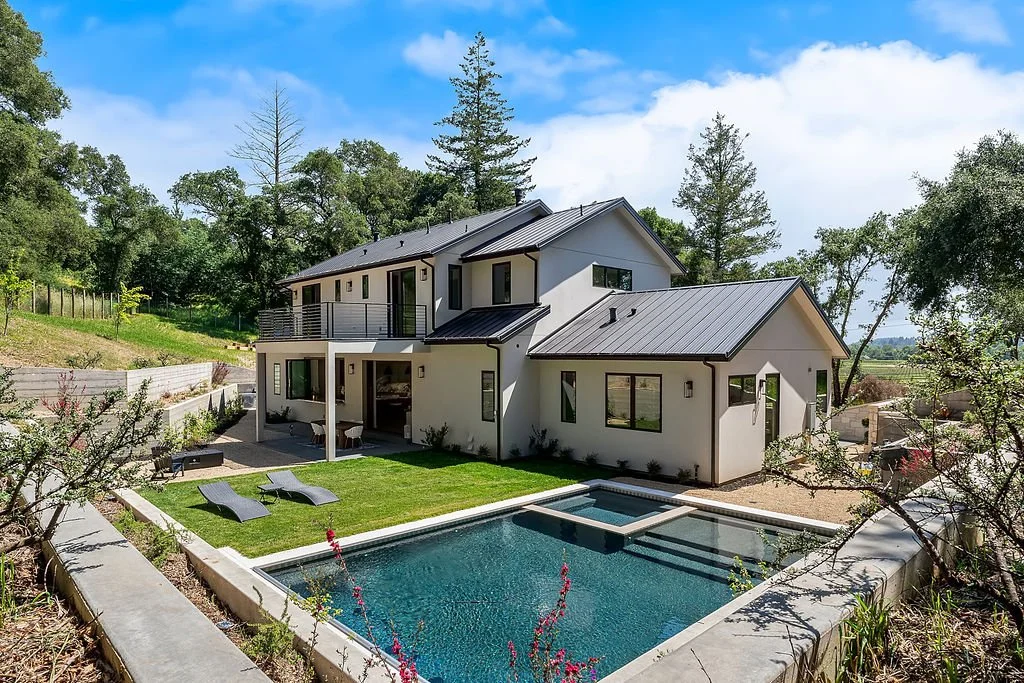 Modern two-story house with a backyard swimming pool, surrounded by trees and greenery under a blue sky.