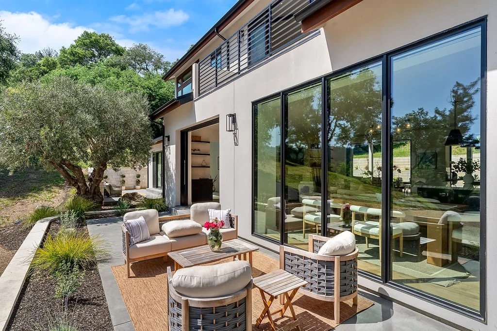 Modern house with large glass sliding doors, outdoor patio furniture, and landscaped yard with trees.