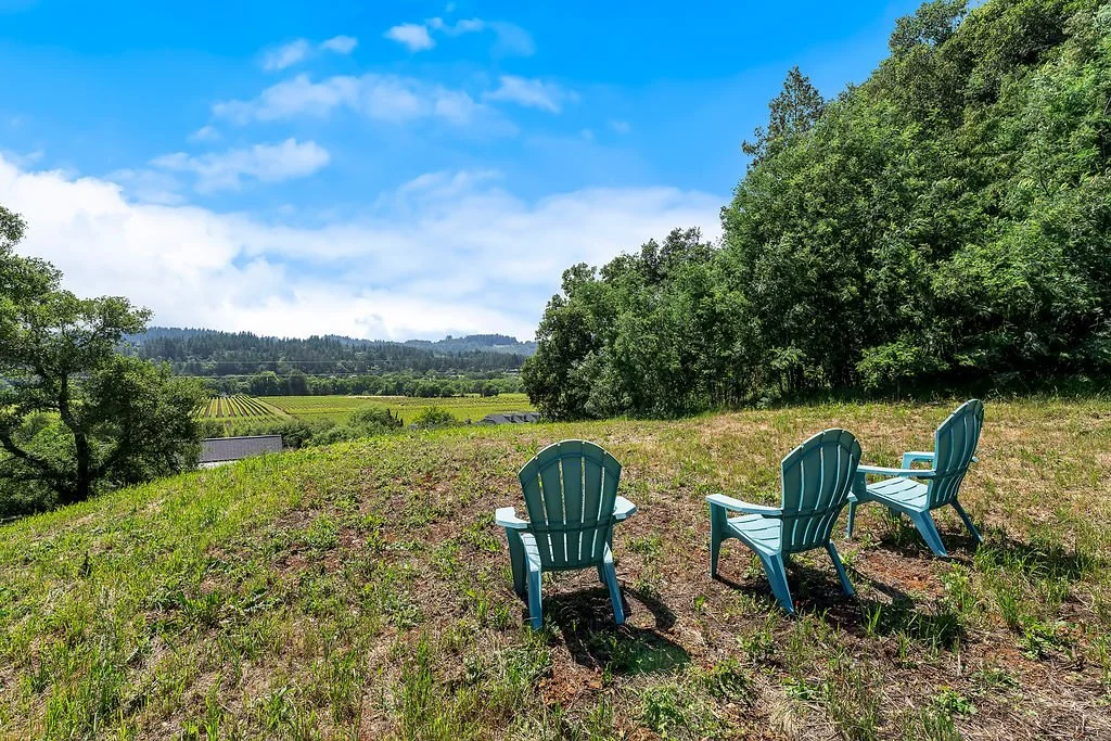 Three blue Adirondack chairs on a grassy hillside overlooking a rural landscape with trees, fields, and distant mountains under a partly cloudy sky.