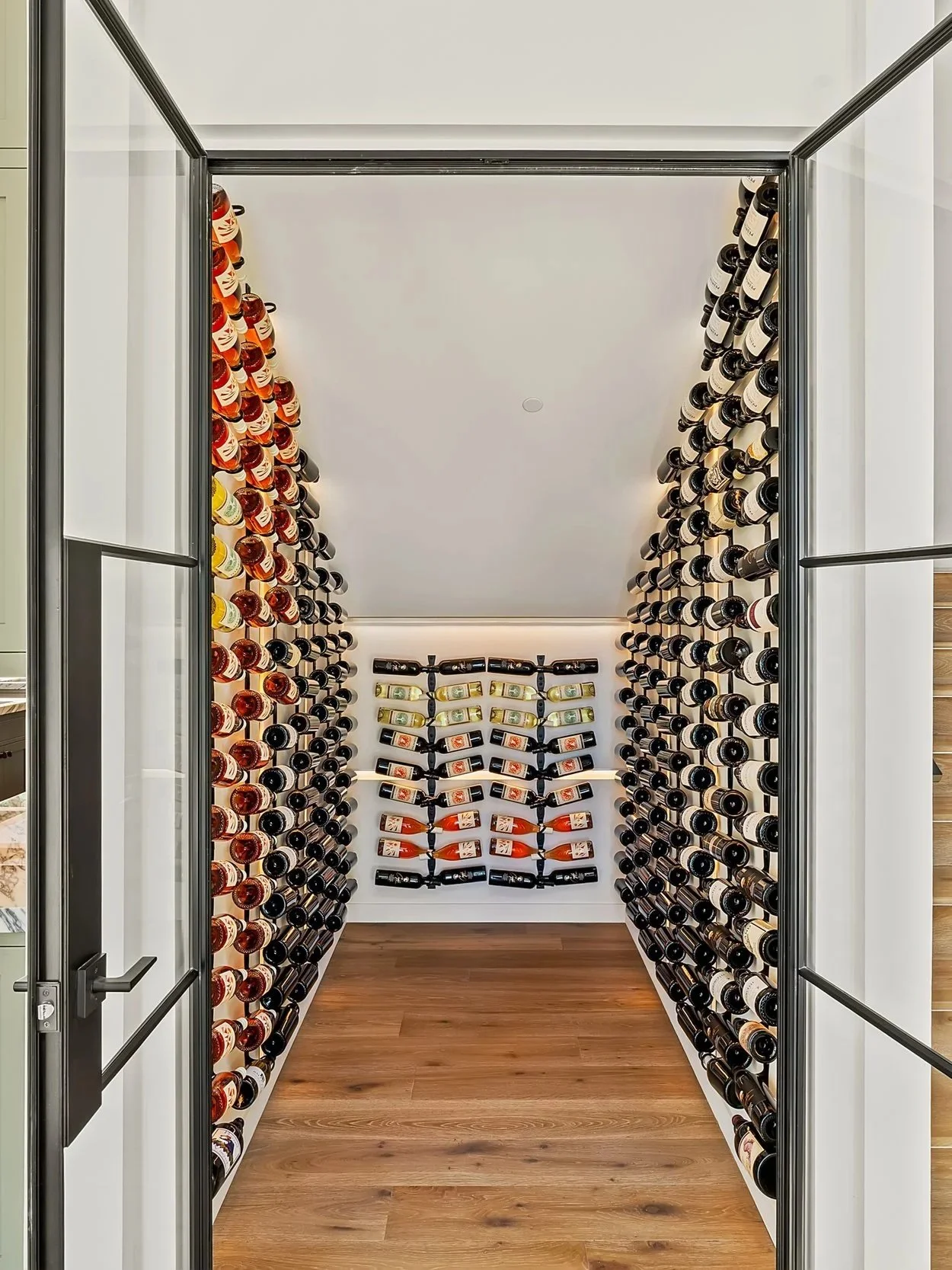 A wine cellar with bottles organized on walls and a white wall with bottles mounted. The cellar has wooden flooring and a glass door.