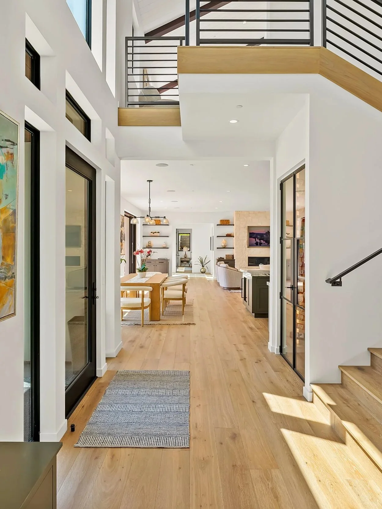 Interior view of a modern, open-concept home with wooden floors, featuring a dining area, living room, and staircase with black railings.