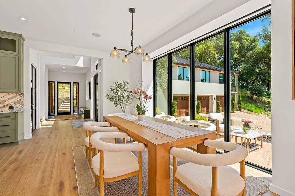 Bright dining area with a wooden table and six cream-colored chairs, large floor-to-ceiling glass windows showing an outdoor patio and a modern house, with trees in the background.