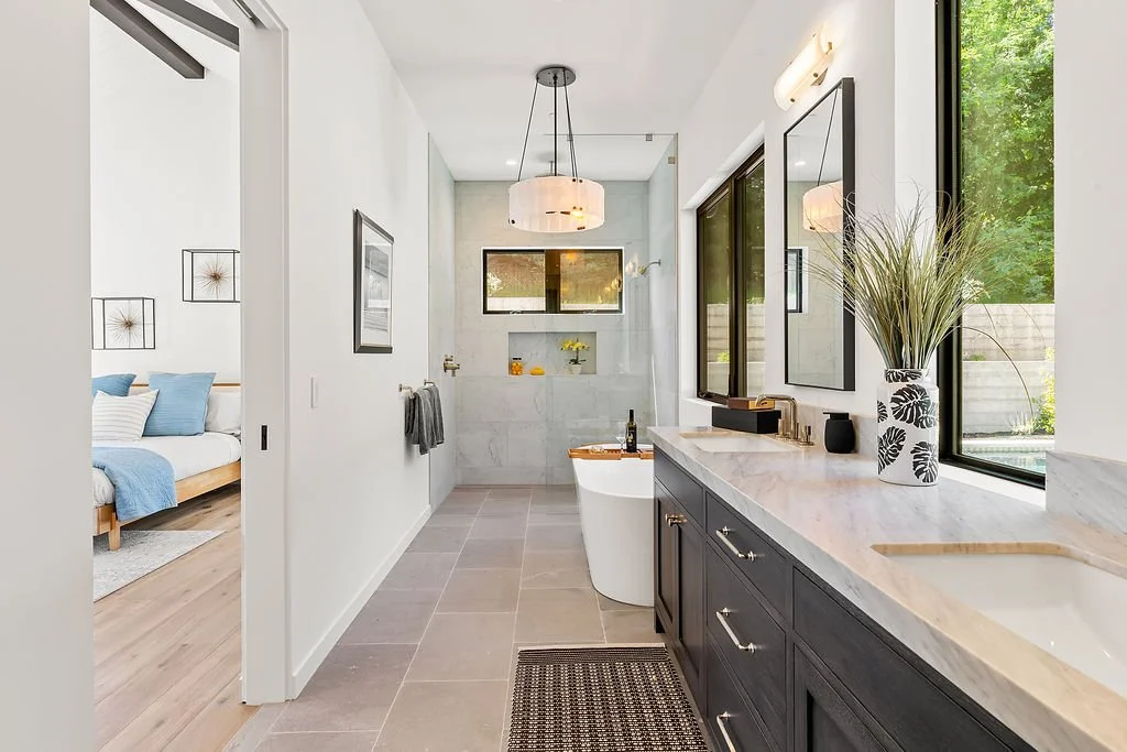 Modern bathroom with a freestanding tub, large window, black vanity with white marble countertop, vases, framed mirrors, and a view into a bedroom with a bed and blue accents.