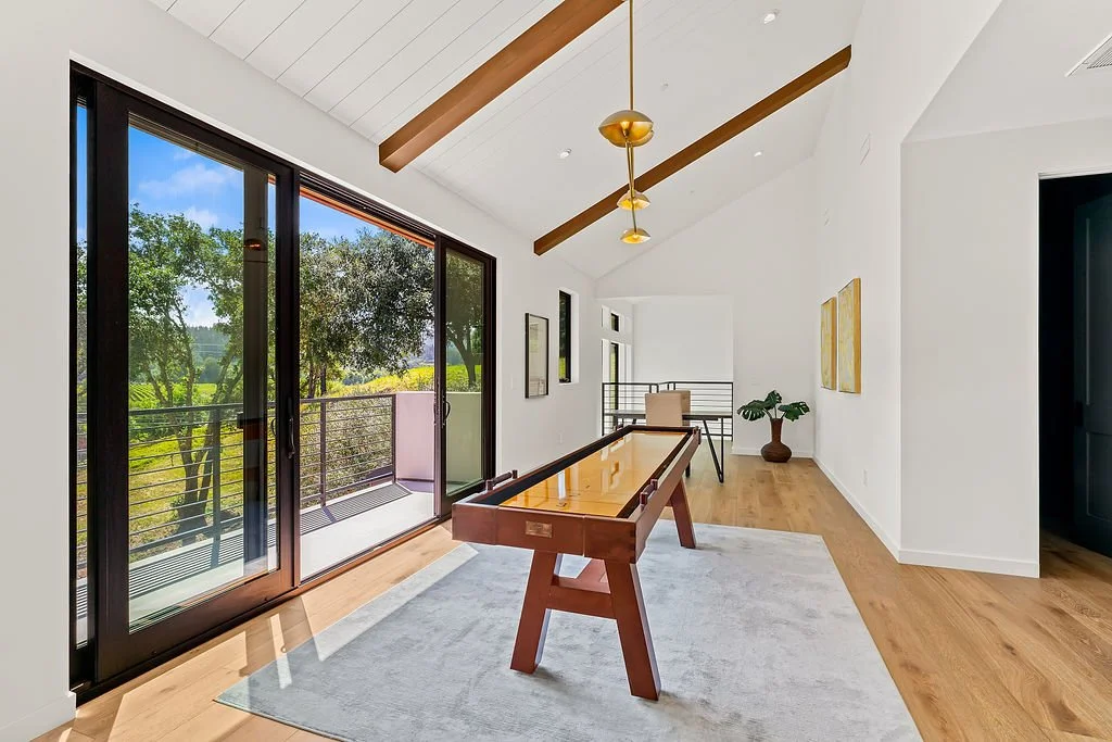 Bright sunroom with large glass sliding doors, a shuffleboard table, a potted plant, and outdoor greenery visible outside.