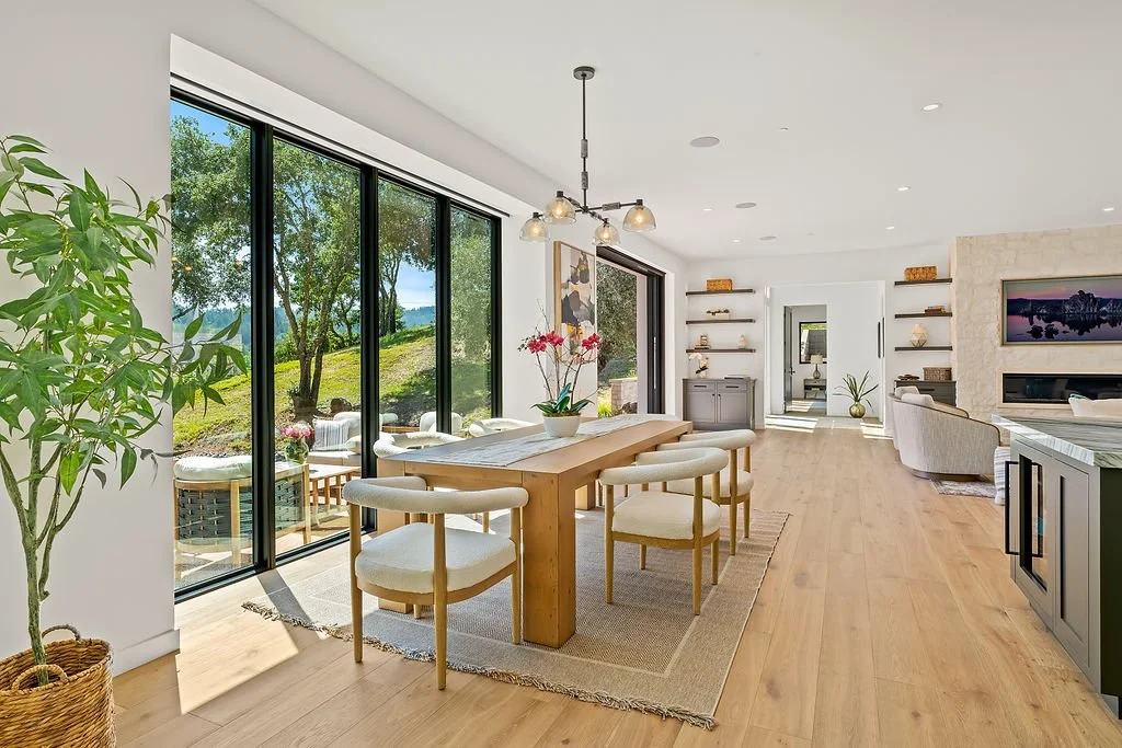 Open-concept living and dining room with large glass sliding doors, wooden flooring, modern furniture, and scenic outdoor view.