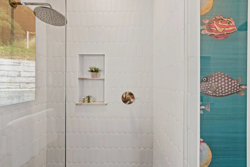 Modern shower with white scalloped tiles, a round showerhead, a small built-in shelf with a potted plant and a small decorative item, and a blue wall with colorful fish illustrations.