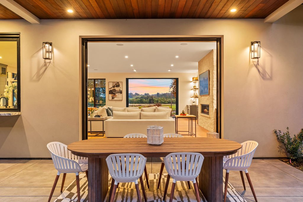 Outdoor patio with a wooden dining table and six white chairs, opening into a living room with a large window, white sofa, and fireplace.