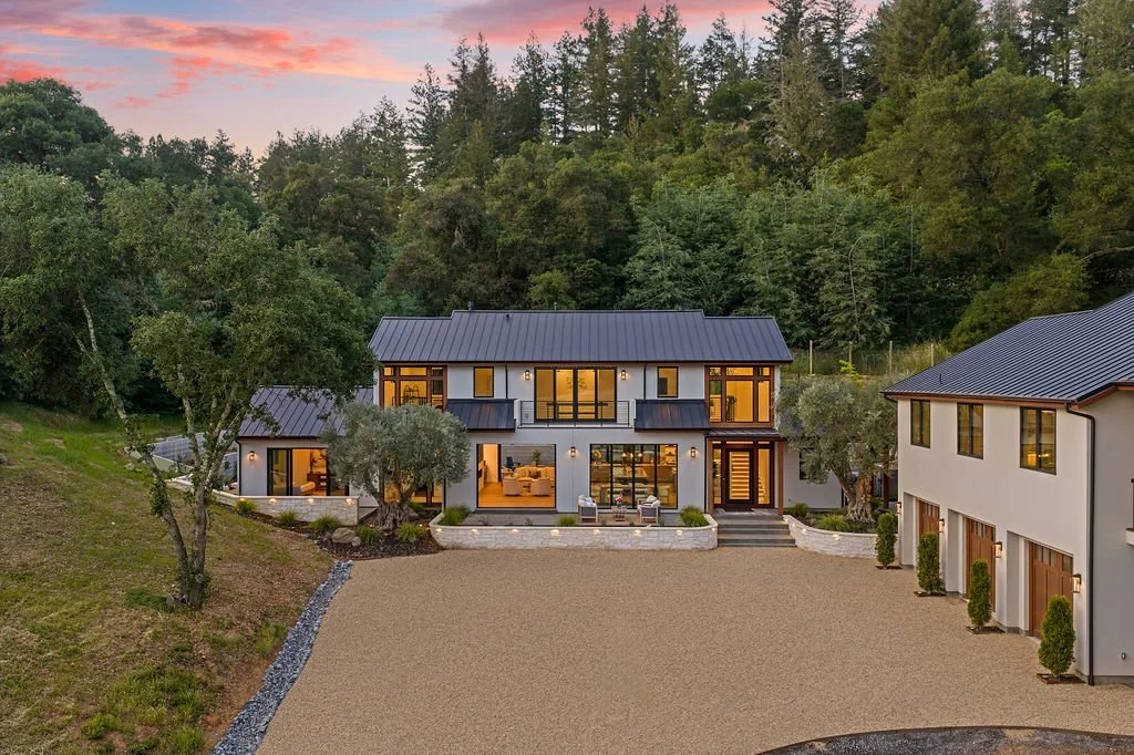 Modern two-story house with large glass windows, surrounded by trees and a gravel driveway, at sunset.