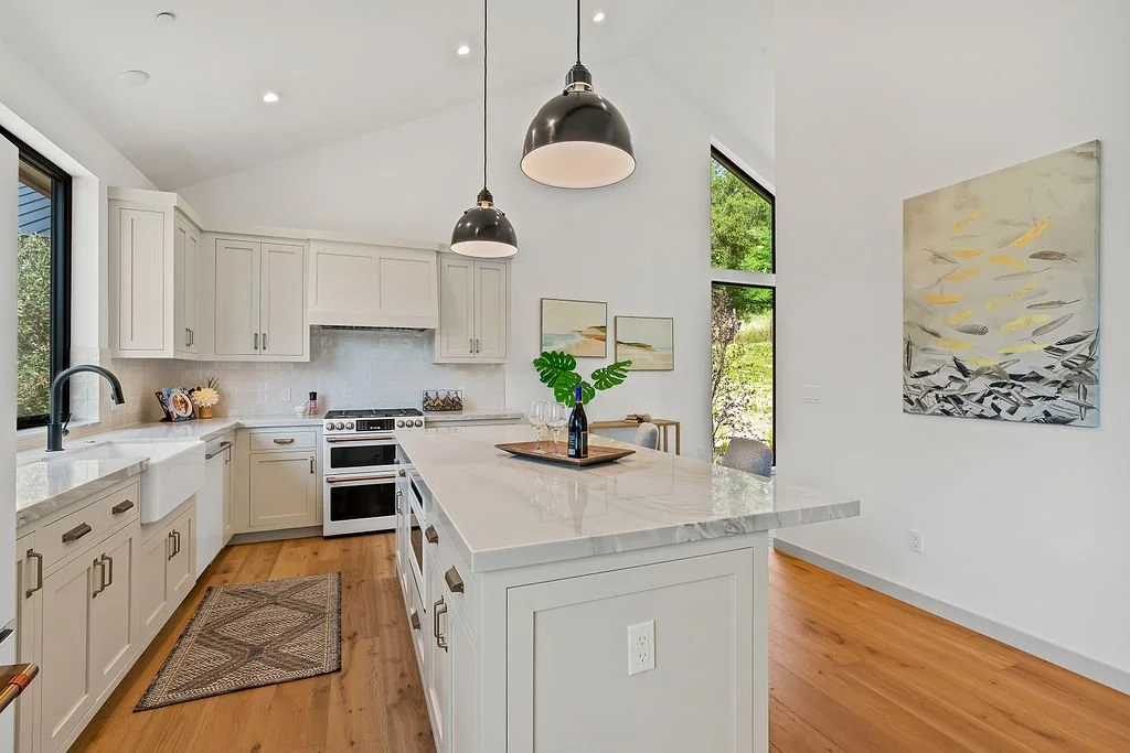 Modern white kitchen with marble island, pendant lights, and large windows showing greenery outside.