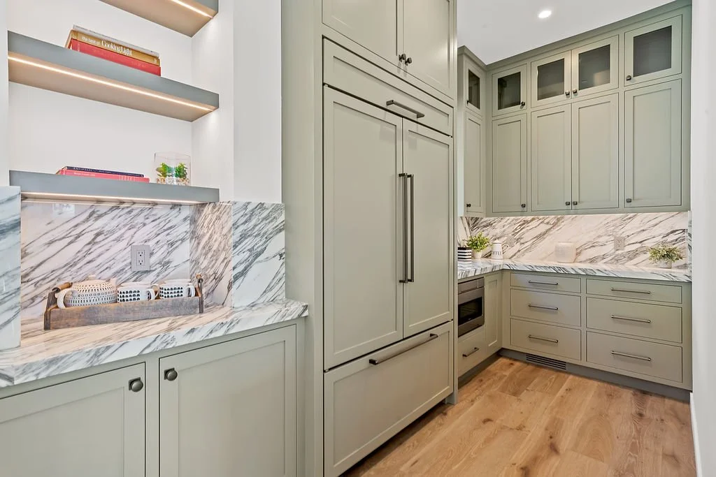 Modern kitchen with sage green cabinets, marble countertops, and wooden flooring.