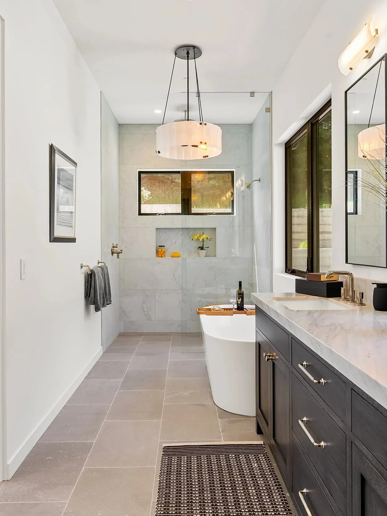 Modern bathroom with white walls, gray floor tiles, a black vanity with silver handles, a marble countertop, a large mirror, and a window showing greenery outside. A white freestanding bathtub with a wooden tray and a wine bottle is visible, along with a built-in shelf with yellow decor and a window in the shower area.