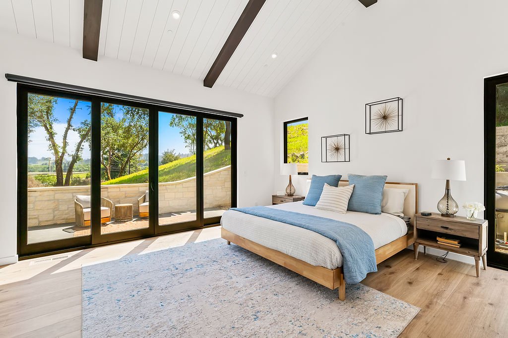 A modern bedroom with a large sliding glass door leading to a balcony, featuring a bed with blue and white bedding, two nightstands with lamps, and wall art, with a view of trees and greenery outside.