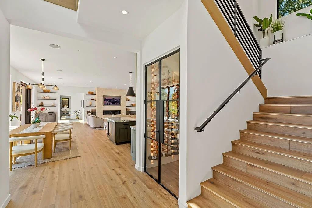 Interior view of a modern home with a wooden staircase, a wine closet, and open living and dining area with shelves, a television, and seating.