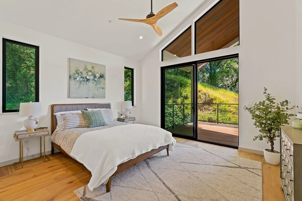 A bright modern bedroom with a large window and glass sliding door leading to a balcony. The room features a bed with white bedding, two nightstands with lamps, and a ceiling fan with wooden blades. There's a plant in a white pot and a piece of artwo