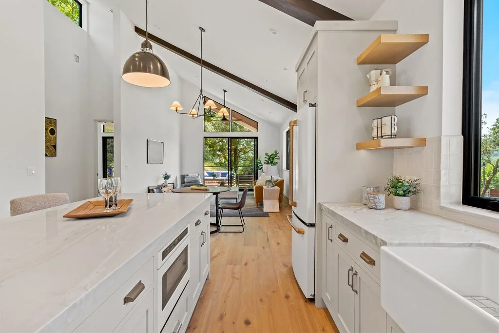 Modern open-concept kitchen with white cabinetry, marble countertops, wooden open shelves, and large windows letting in natural light.
