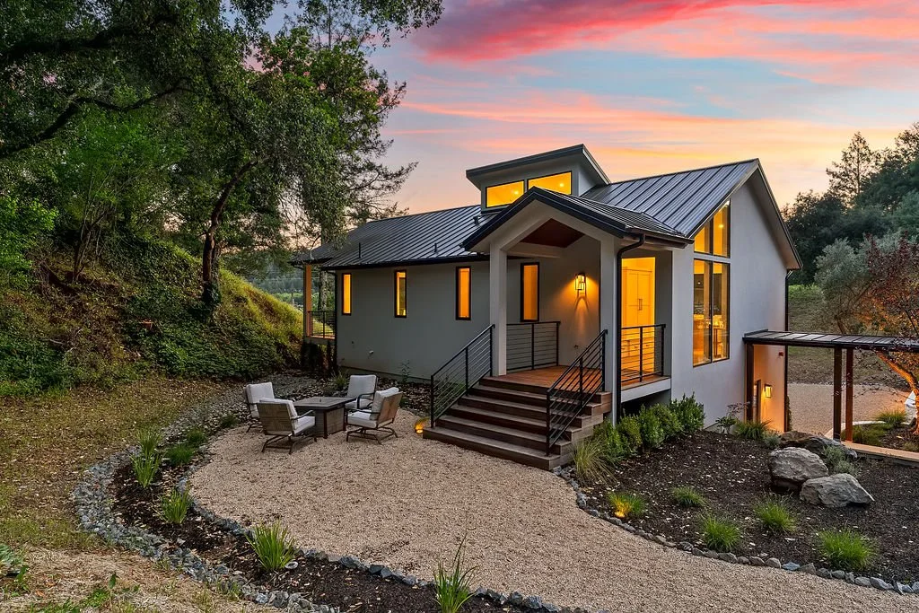 Modern white house with metal roof, large windows, and front porch with stairs, surrounded by trees and landscaped yard during sunset.