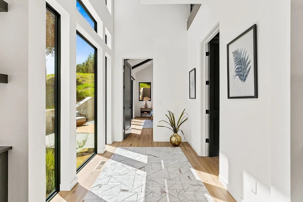 Bright hallway with large windows, white walls, wooden flooring, framed artwork, and a potted plant in a gold vase.