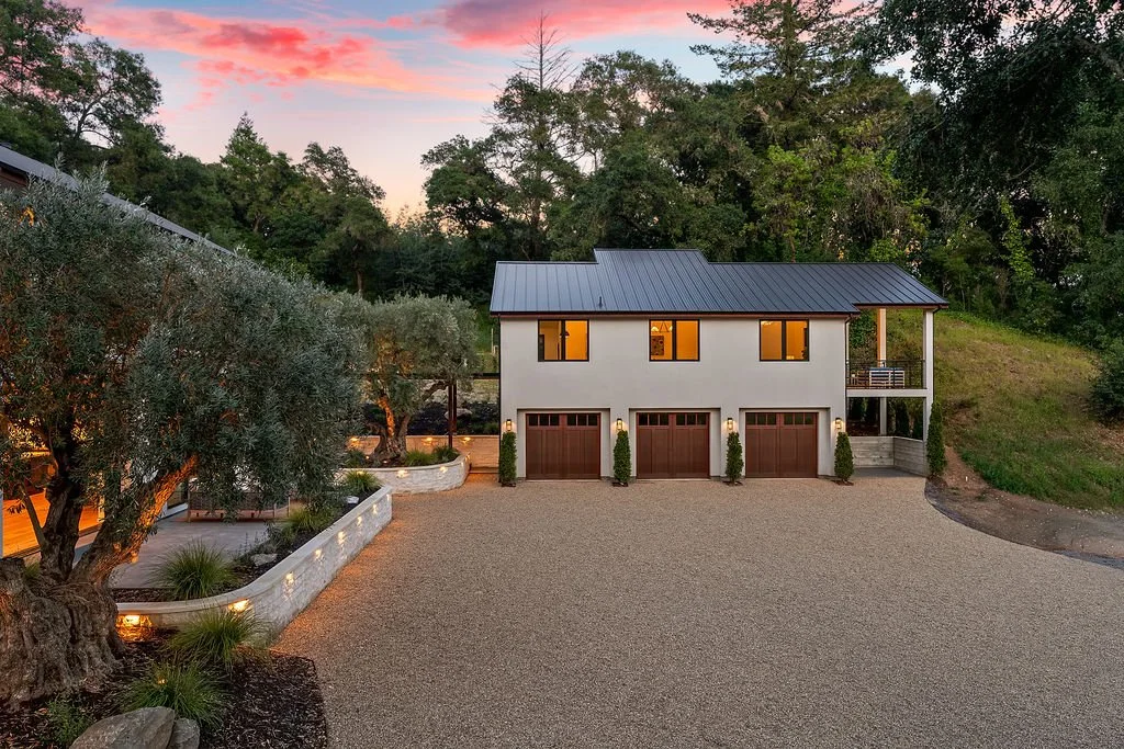 A modern two-story house with a four-car garage, situated on a gravel driveway and surrounded by trees, during sunset.