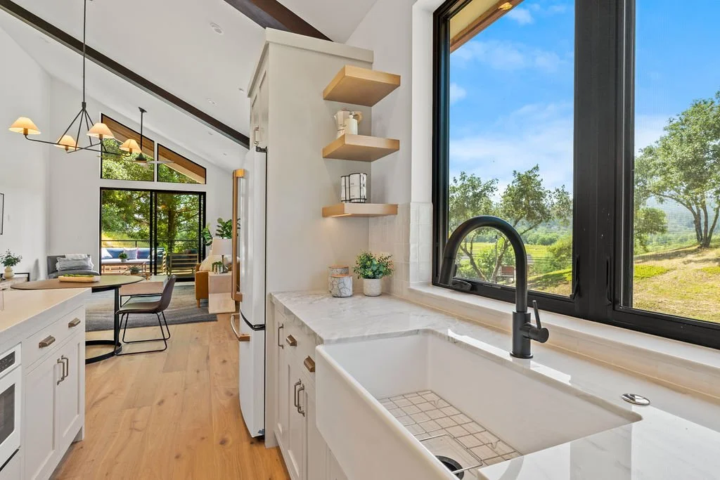View of a bright kitchen with a window overlooking green trees. The kitchen has white cabinets, a black faucet over a farmhouse sink, and wooden open shelves. The adjacent dining and living area features a dining table, chairs, and large windows with