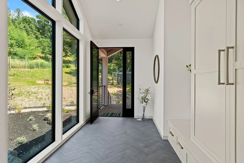 Bright entryway with large floor-to-ceiling windows, a glass door leading outside to a deck, white cabinets, and a vase with white flowers.
