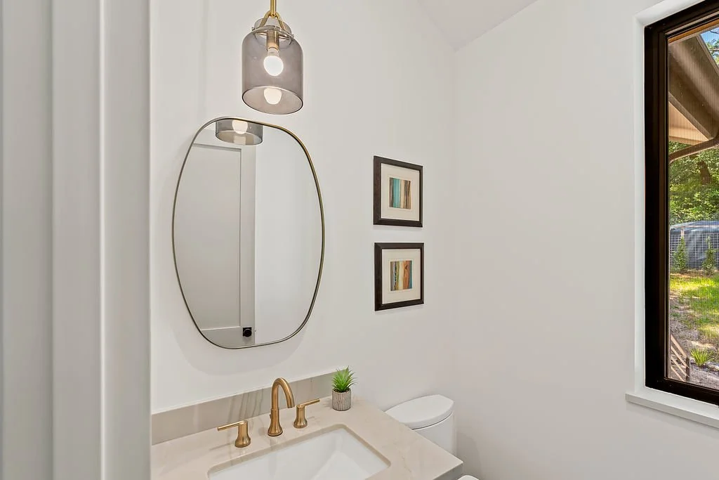 Modern bathroom with white walls, oval mirror, brass fixtures, small plant, framed artwork, toilet, and a large window showing outdoors.