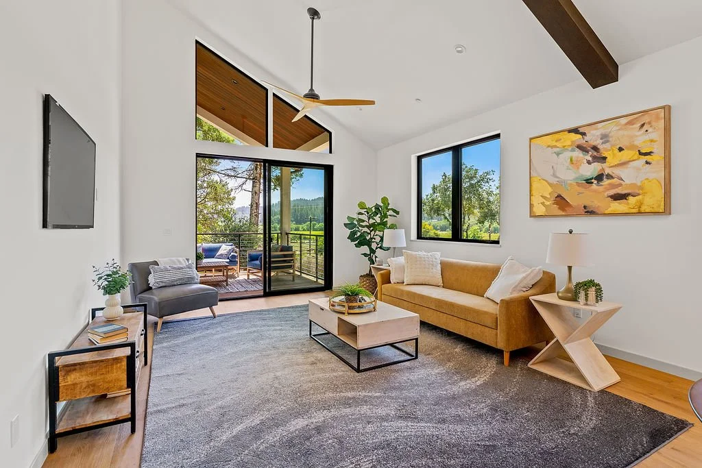 Modern living room with a sofa, coffee table, side table, and a balcony with outdoor seating overlooking greenery.