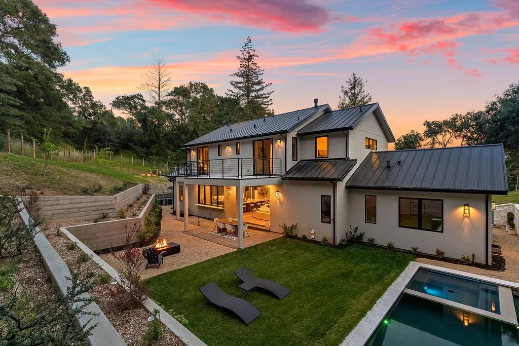 Modern two-story house with large windows, outdoor patio with dining area and fire pit, green lawn with lounge chairs, a swimming pool, and surrounding trees at sunset.