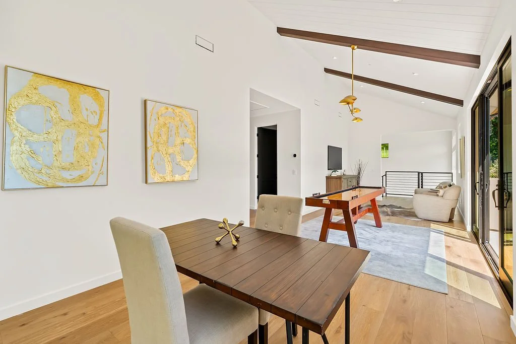 Modern open-concept living and dining area with white walls, hardwood floors, abstract gold and white artwork, a dining table with beige chairs, a foosball table, a lounge chair, a television, and large sliding glass doors leading to an outdoor space