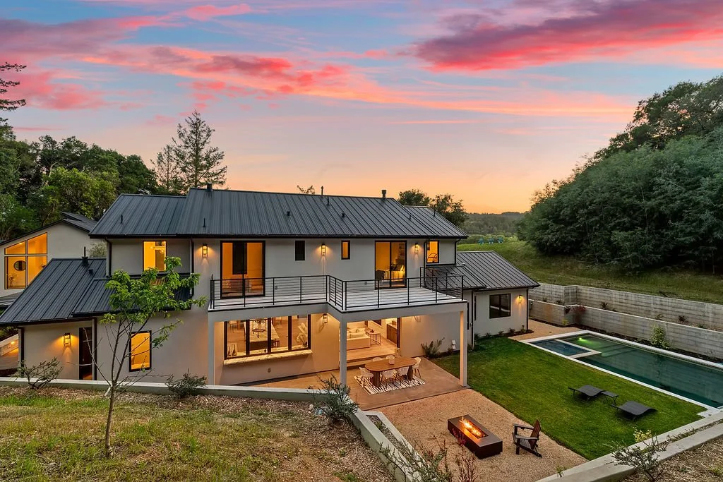 Modern two-story house with a dark metal roof, large windows, and a spacious balcony, overlooking a backyard with a swimming pool, patio furniture, and a fire pit during sunset.