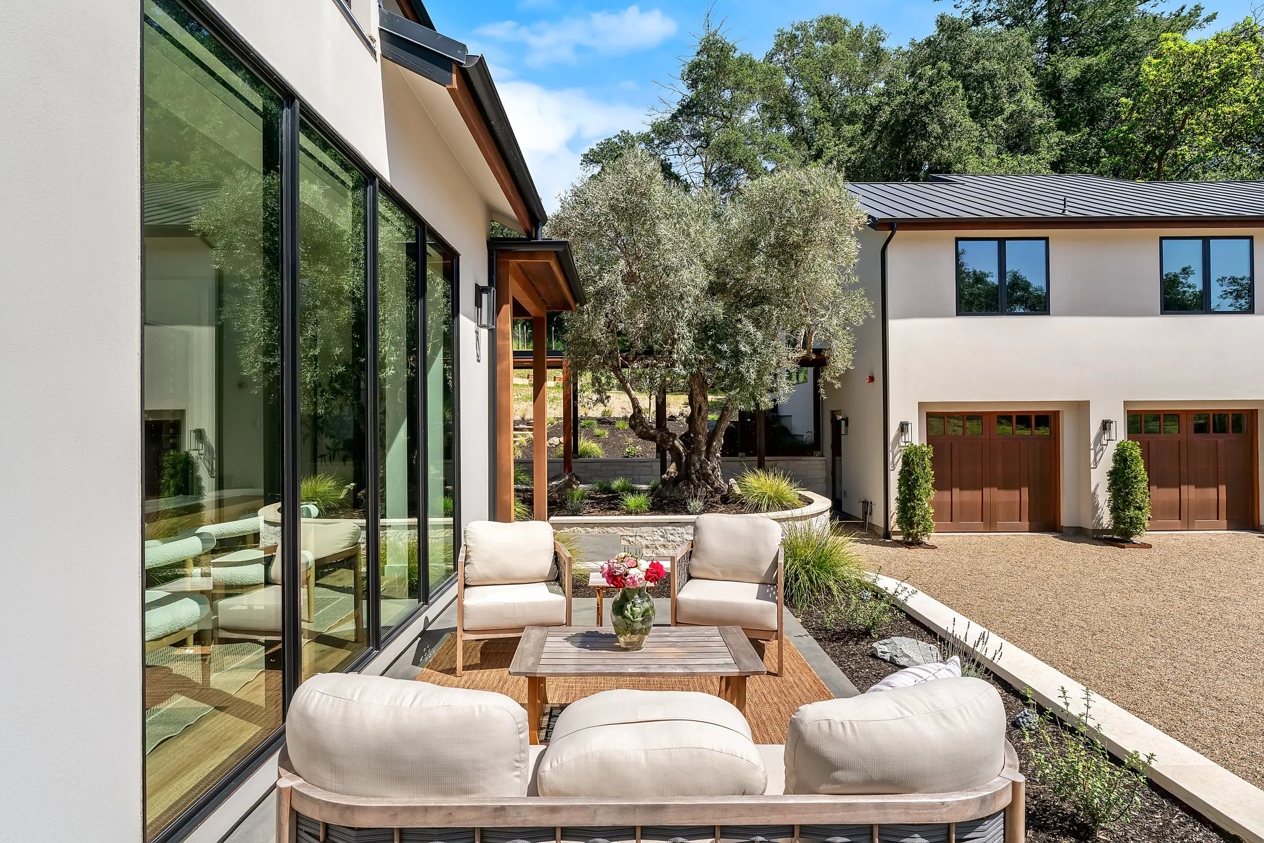 Modern house with large glass windows, outdoor seating area with beige cushions, wooden table with flowers, trees, and neighboring house with gray roof.