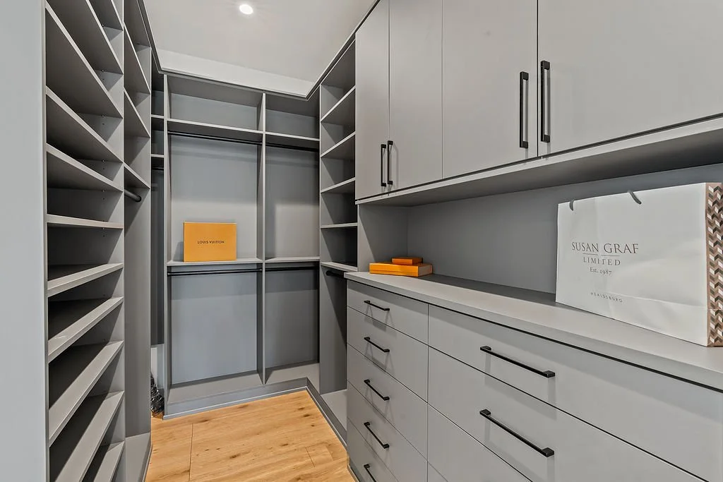 Walk-in closet with gray shelves and drawers, some with black handles, and a few orange boxes on the shelves. Floor is wooden, and the closet is well-lit.