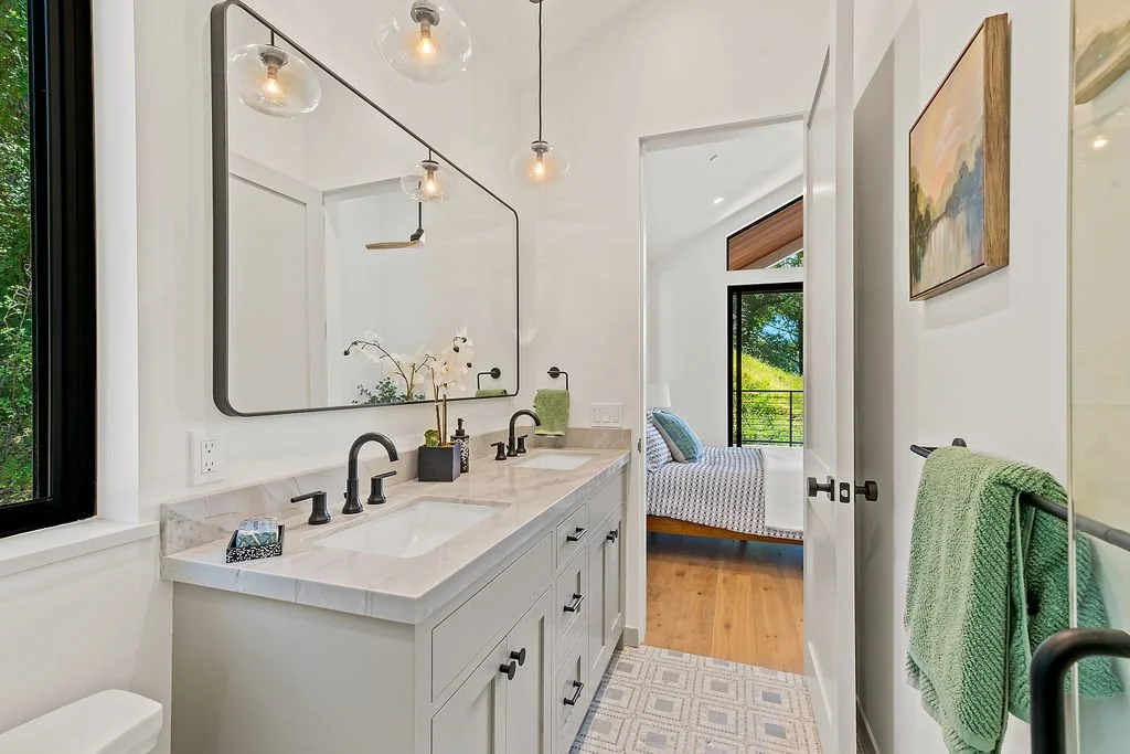 Bathroom with a double vanity, large mirror, black fixtures, and a window showing greenery outside. A bedroom with a bed, pillows, large window, and wooden flooring is visible through an open door.