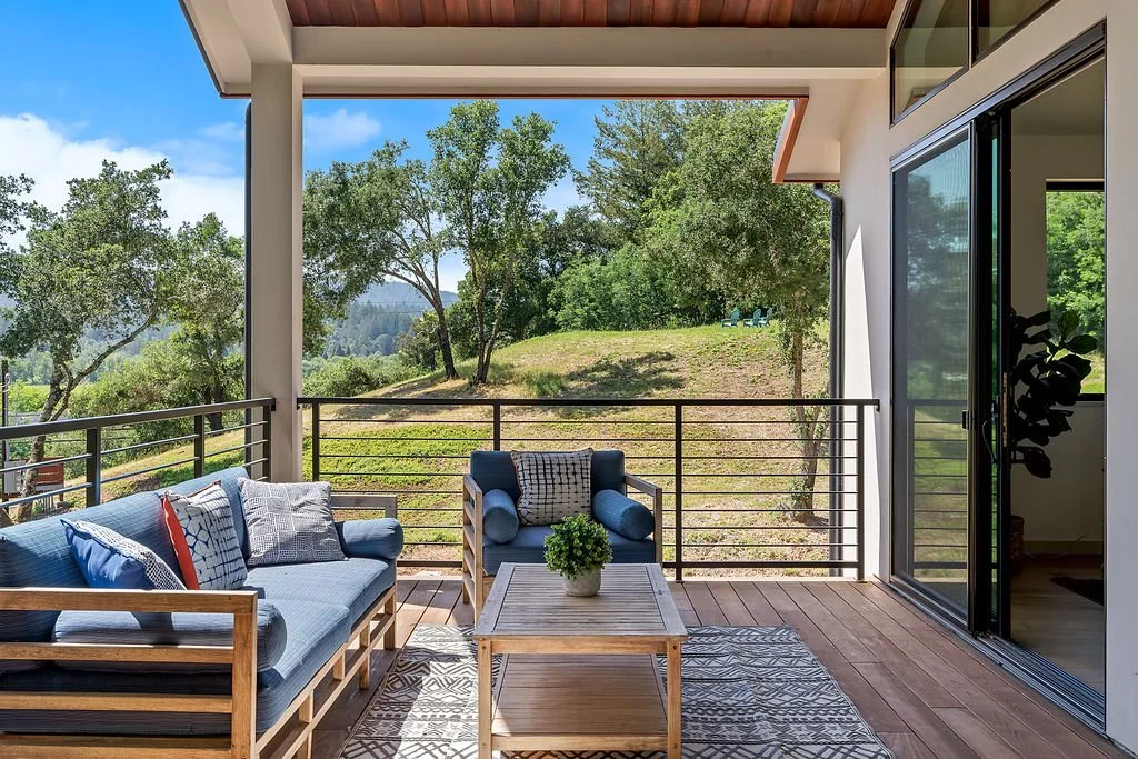 Outdoor balcony with wooden furniture and a view of trees and grassy hillside.