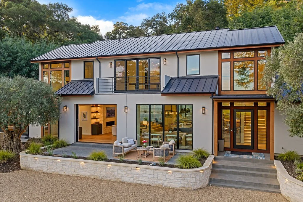 Modern two-story house with large windows and a metal roof, surrounded by trees and landscaped yard.