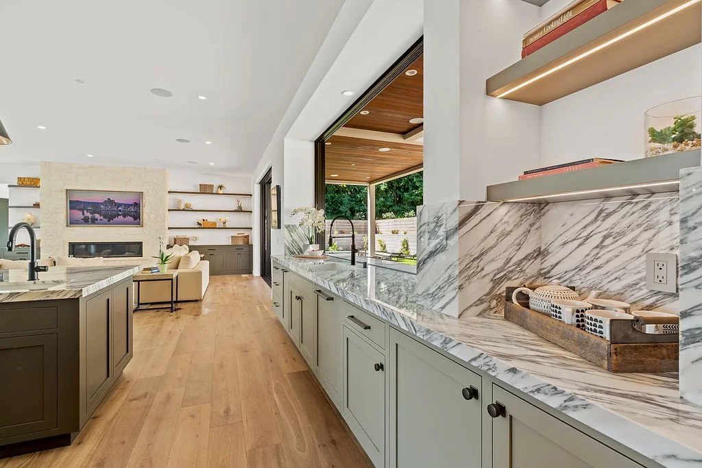 Modern kitchen with white cabinets, marble countertops, and open shelves. Large window overlooking a patio and backyard. Bright interior with natural light.