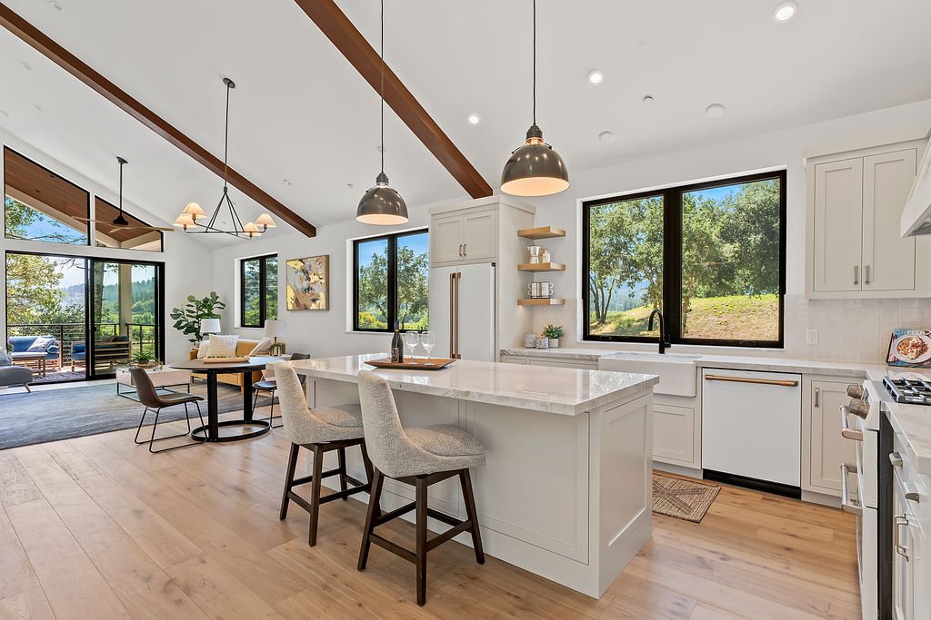 Modern open-concept kitchen with white cabinetry, a large island, black pendant lights, and large windows bringing in natural light, with a view of greenery outside.