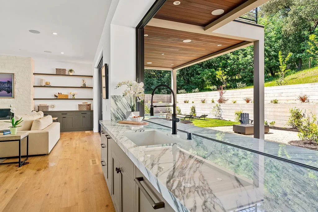 Open kitchen with marble countertops overlooking a backyard patio with trees and shrubs.
