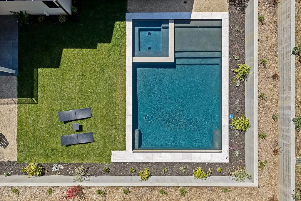 An aerial view of a backyard pool area with two pool loungers, a small table, a grassy lawn, and plants bordering the patio.