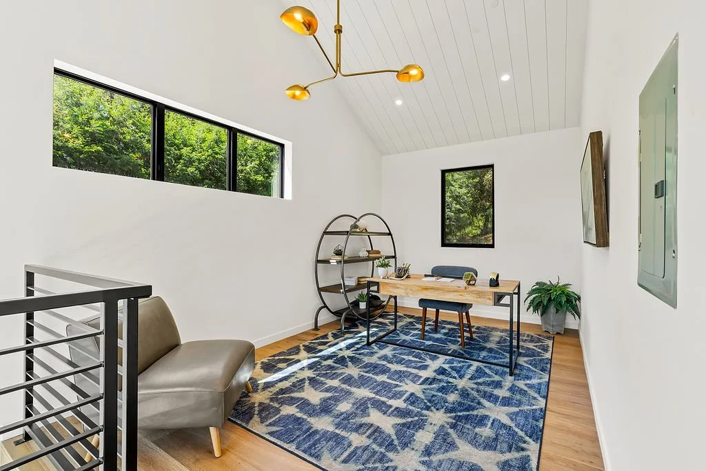 Modern home office with a wooden desk, black chair, blue patterned rug, and large windows showing green trees outside.