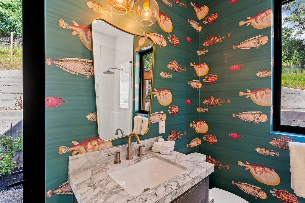 Bathroom with fish-themed wallpaper featuring various colorful fish on teal background, marble countertop, rectangular mirror, brass faucet, and a window showing greenery outside.