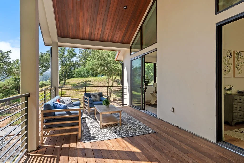 Outdoor balcony with wooden flooring, railing, and furniture including a sofa and armchair with cushions, overlooking a green landscape with trees and grass.